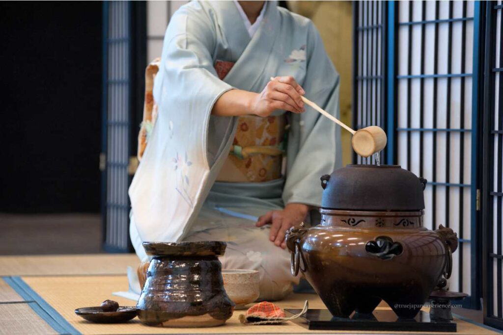 A woman in a light blue kimono performing a Japanese tea ceremony, pouring hot water with a bamboo ladle into a traditional iron kettle.