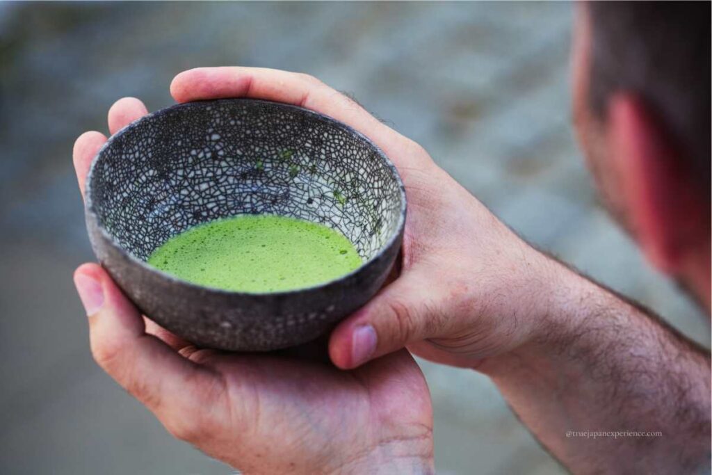A close-up of hands holding a textured ceramic bowl filled with freshly whisked matcha during a Japanese tea ceremony.