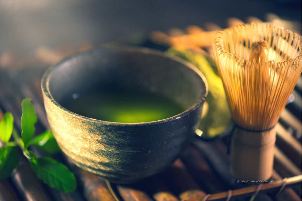A rustic ceramic bowl filled with matcha green tea beside a bamboo whisk (chasen), set on a wooden surface with natural lighting.