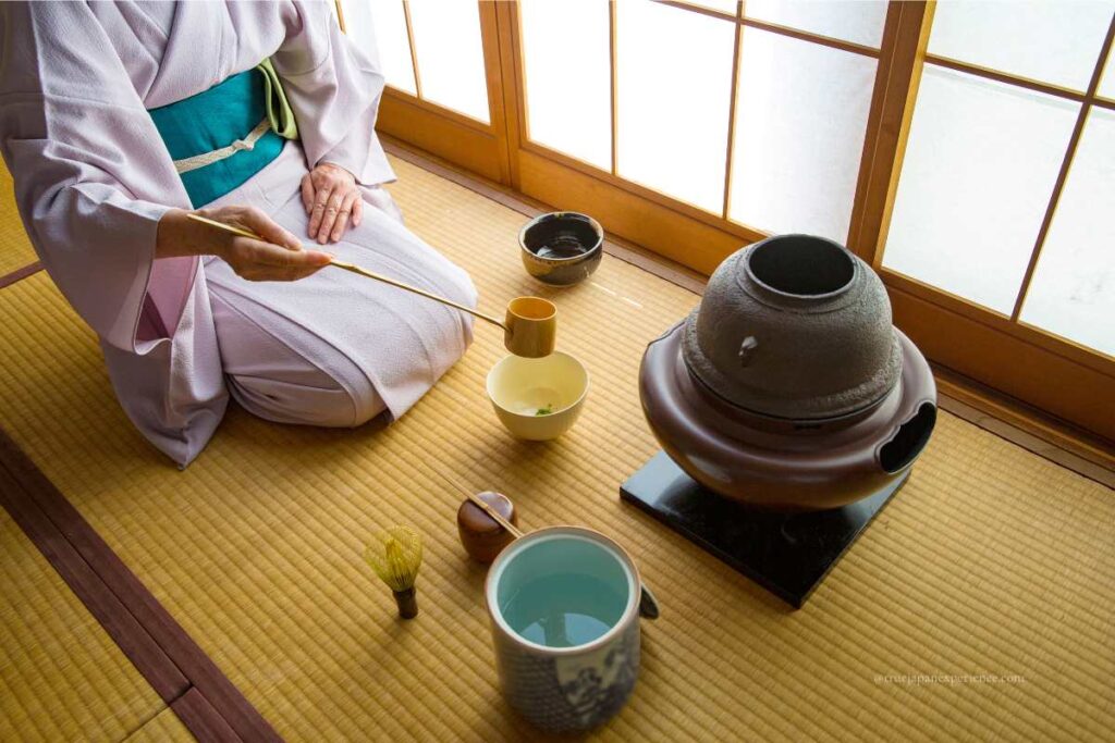 A person in a kimono preparing tea during a Japanese tea ceremony, using a bamboo ladle over a traditional iron kettle on a tatami mat.