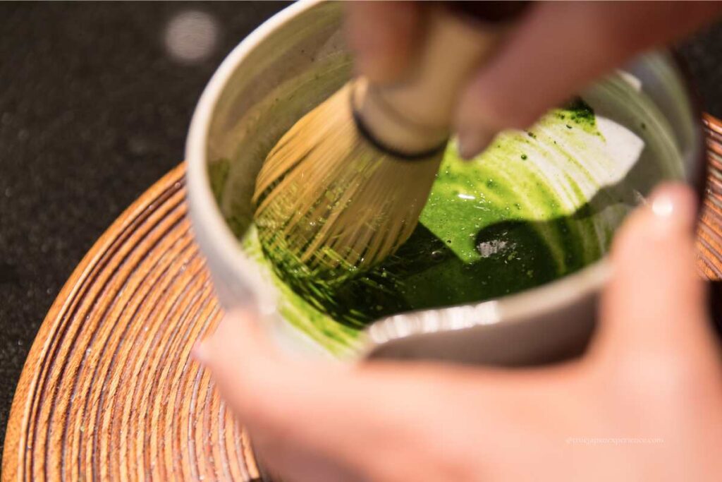 Close-up of hands whisking matcha in a tea bowl with a bamboo whisk (chasen) during a Japanese tea ceremony.