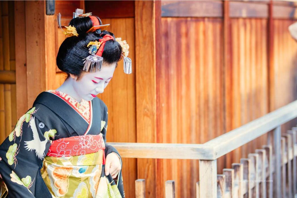 A maiko in traditional makeup and ornate kimono walking past a wooden building in Kyoto.