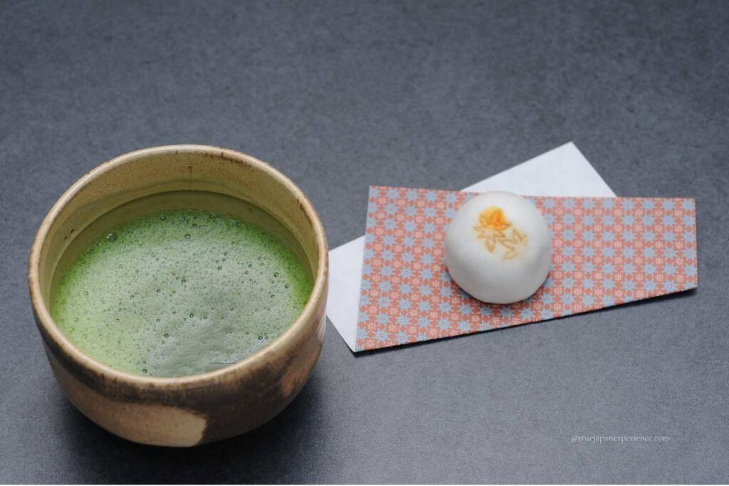 A bowl of frothy matcha green tea served with a Japanese sweet (wagashi) on patterned paper, part of a traditional tea ceremony.