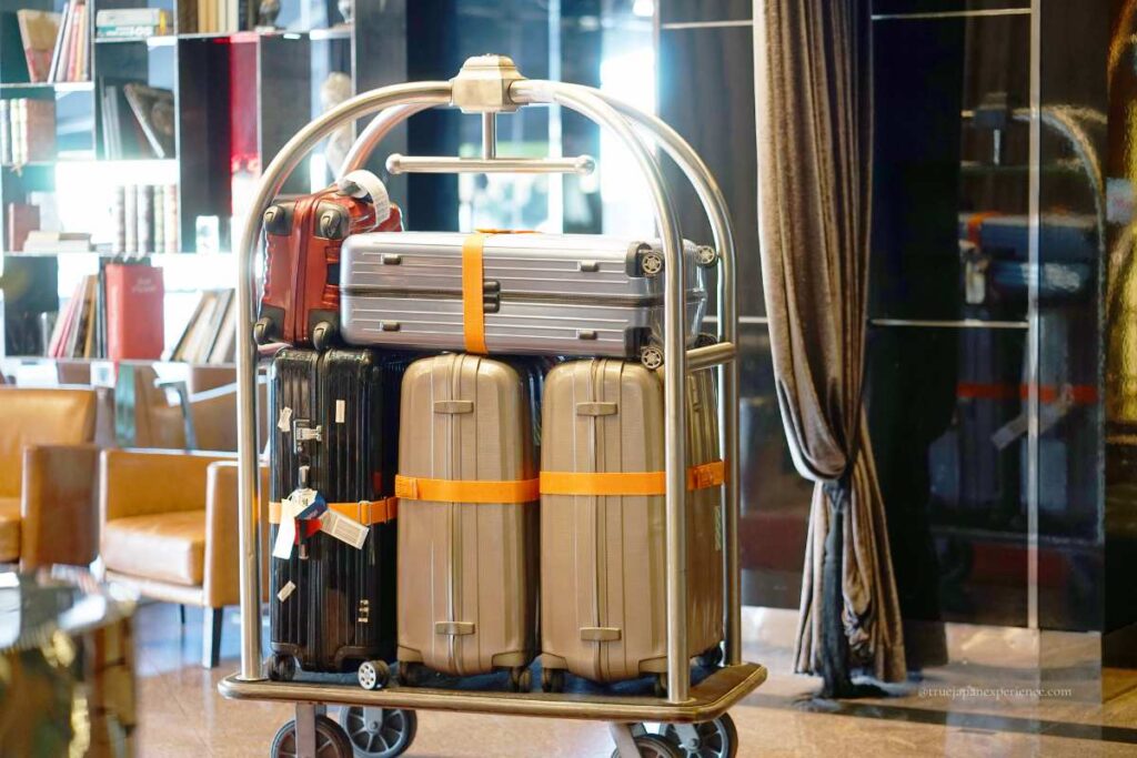 A hotel luggage cart loaded with several suitcases in various colors, secured with orange straps, in a modern hotel lobby.