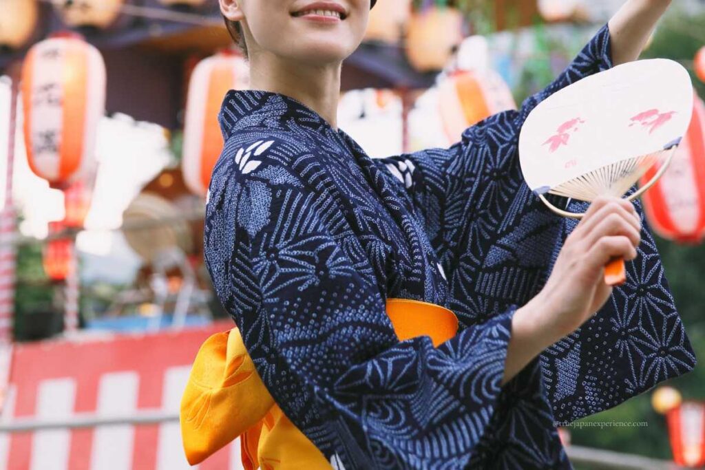 Woman wearing a blue yukata and holding a fan at a Japanese summer festival.
