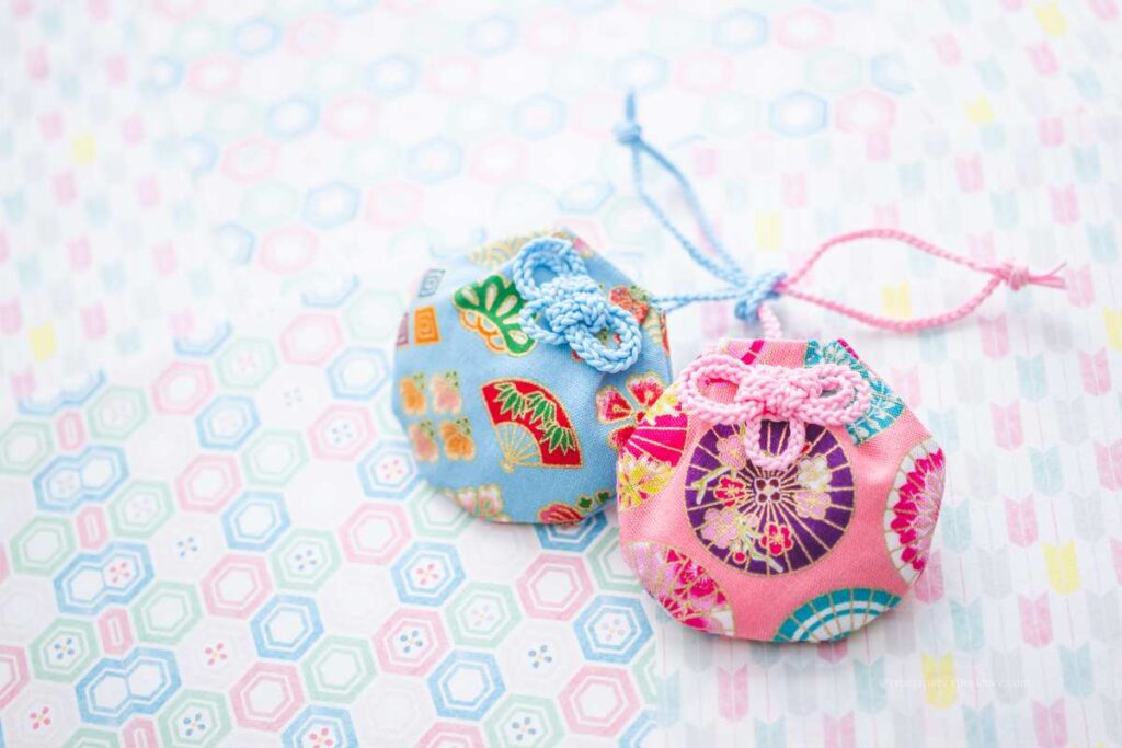 Two round Japanese omamori made with colorful kimono-style fabric, displayed on a pastel patterned background.