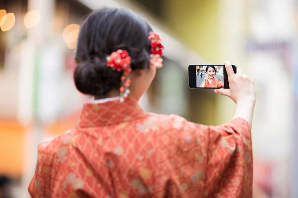 Woman in kimono smiling at her phone while taking a selfie on a scenic street