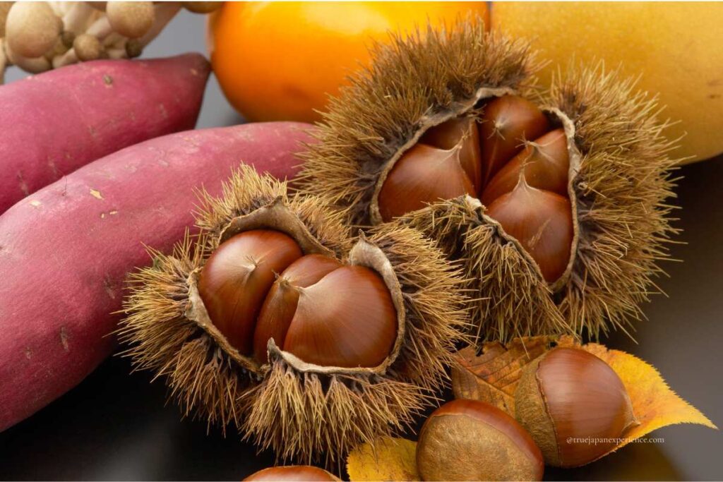 Close-up of Japanese autumn foods including spiky chestnut shells, sweet potatoes, mushrooms, and a persimmon, symbolizing seasonal harvest.