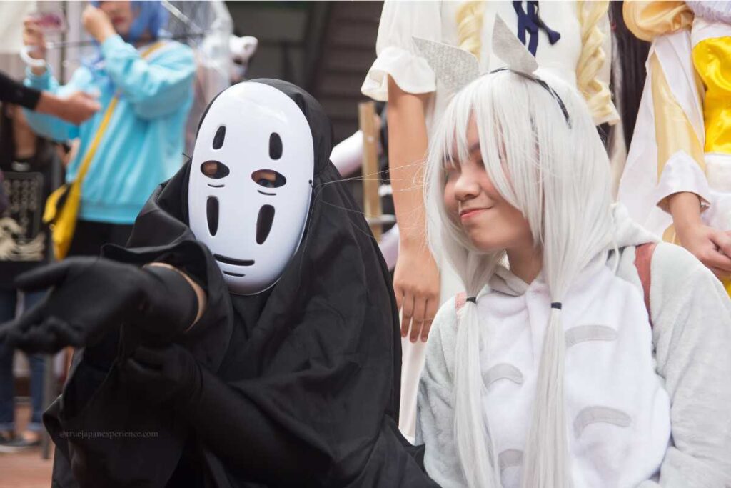 Two people in anime character costumes at a cosplay event in Japan, one dressed as No-Face from Spirited Away and the other as Totoro with white hair and animal ears.