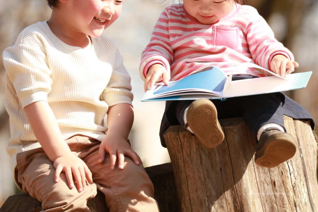 Two young children sitting on tree stumps outdoors, smiling and reading a picture book together on a sunny autumn day.