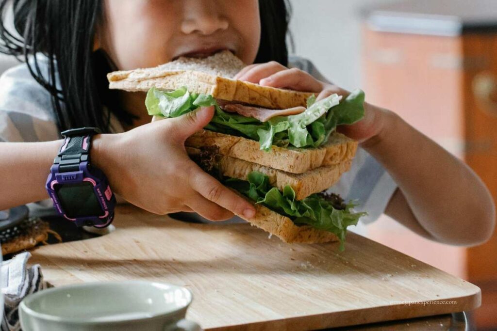A child holding and biting into a giant sandwich stacked with layers of bread, lettuce, and ham, wearing a purple digital watch.