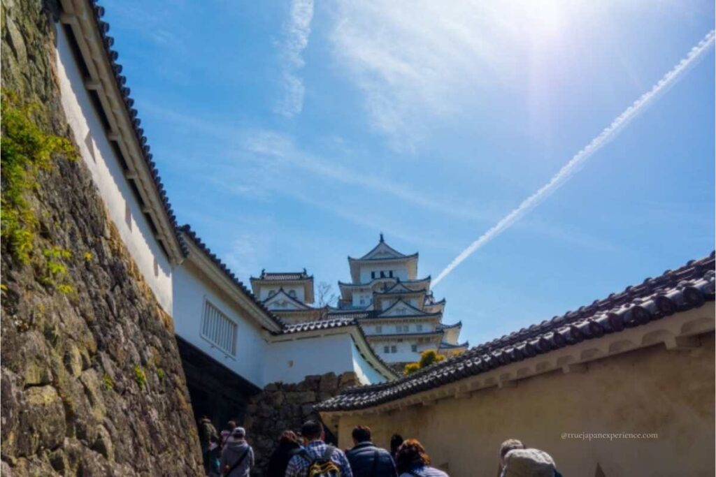 Visitors walking through stone and white-walled corridors toward the main keep of Himeji Castle under a sunny sky with a jet trail