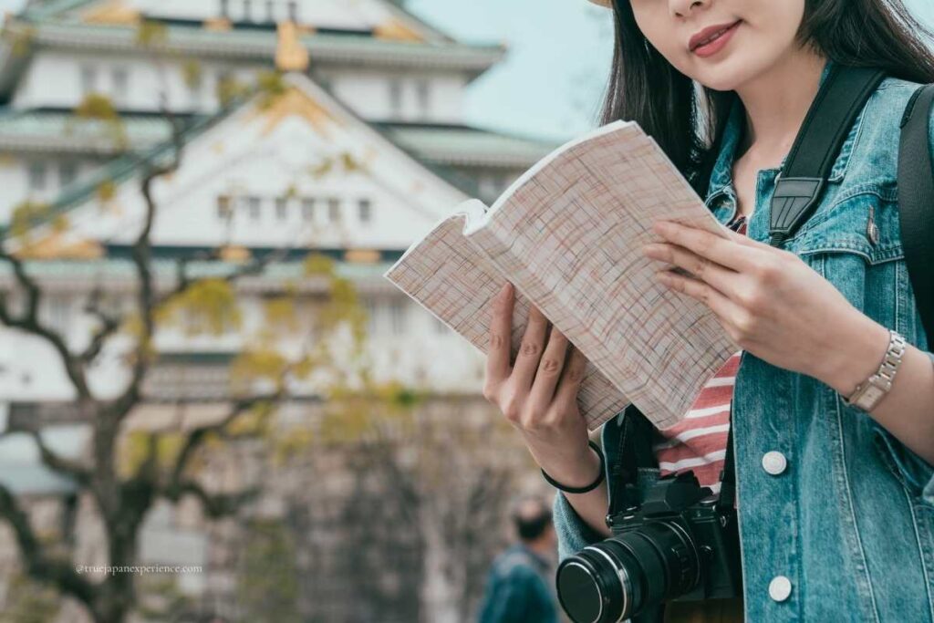 Female tourist holding a map and wearing a camera around her neck, standing in front of a Japanese castle