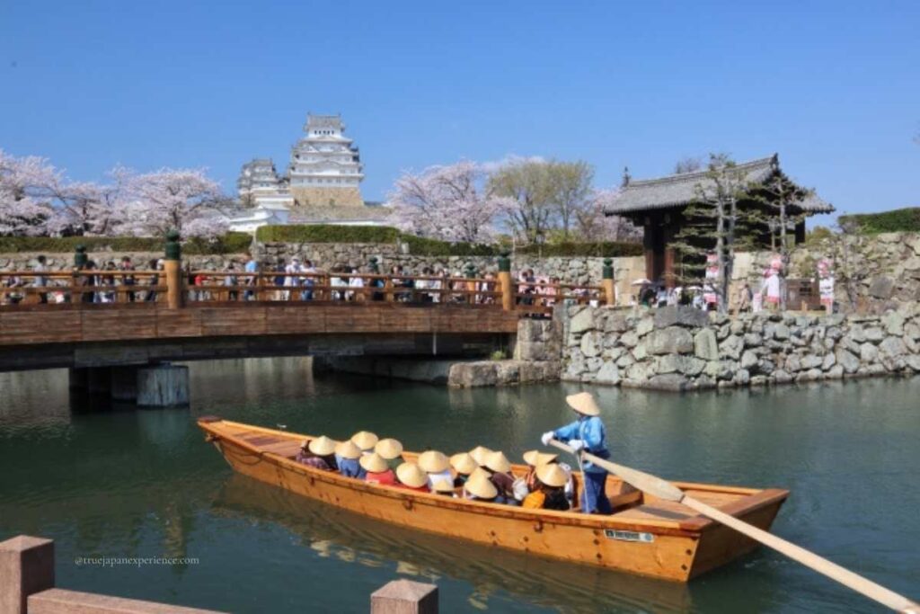 Tourists riding a traditional Japanese boat in the moat of Himeji Castle, with cherry blossoms, a wooden bridge, and stone walls in the background