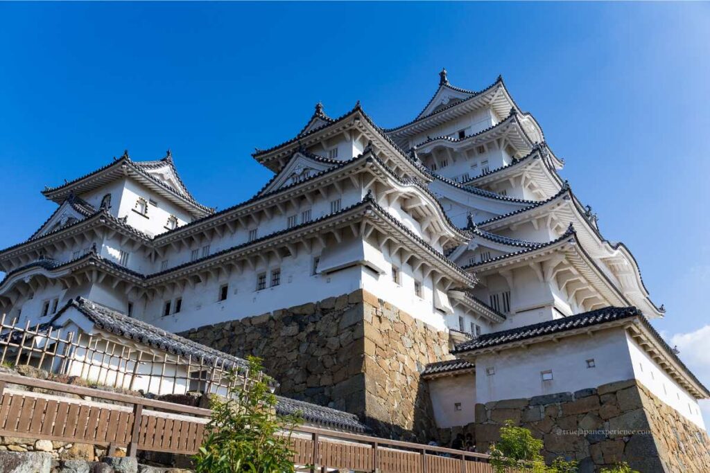 Close-up view of the main keep of Himeji Castle from below, showing its white walls, stone base, and layered rooftops under a clear blue sky