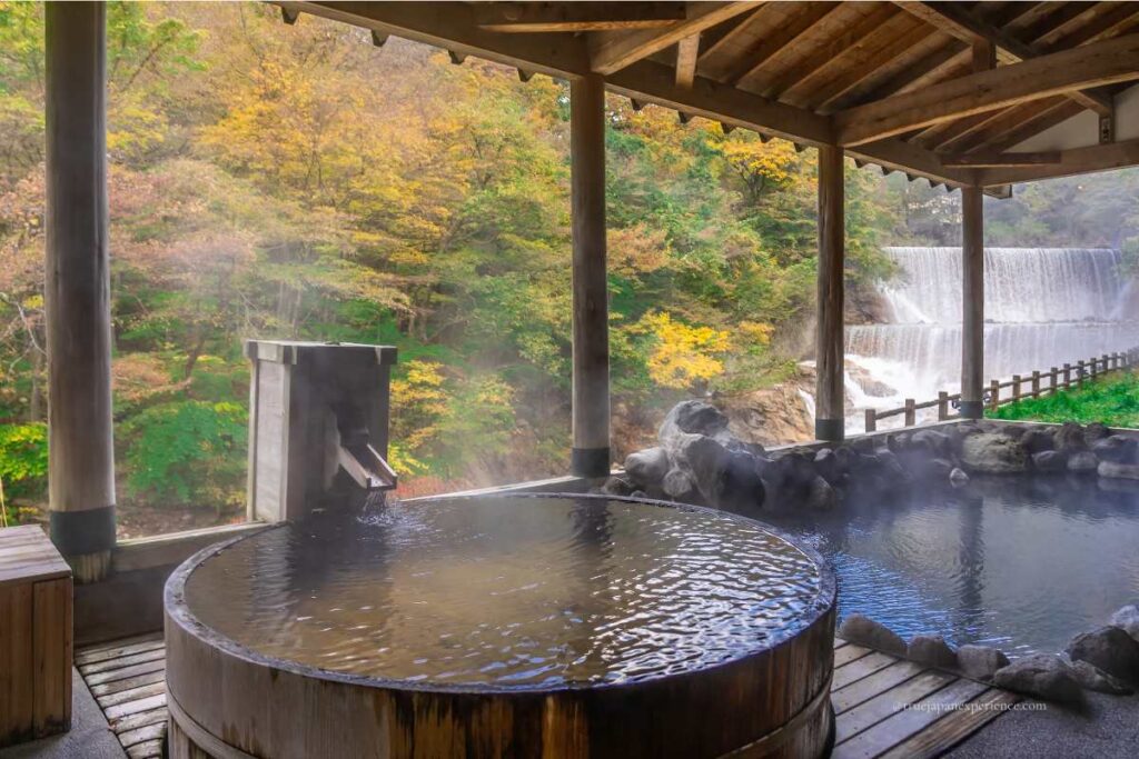 Outdoor hot spring bath with a wooden tub and a waterfall view in autumn