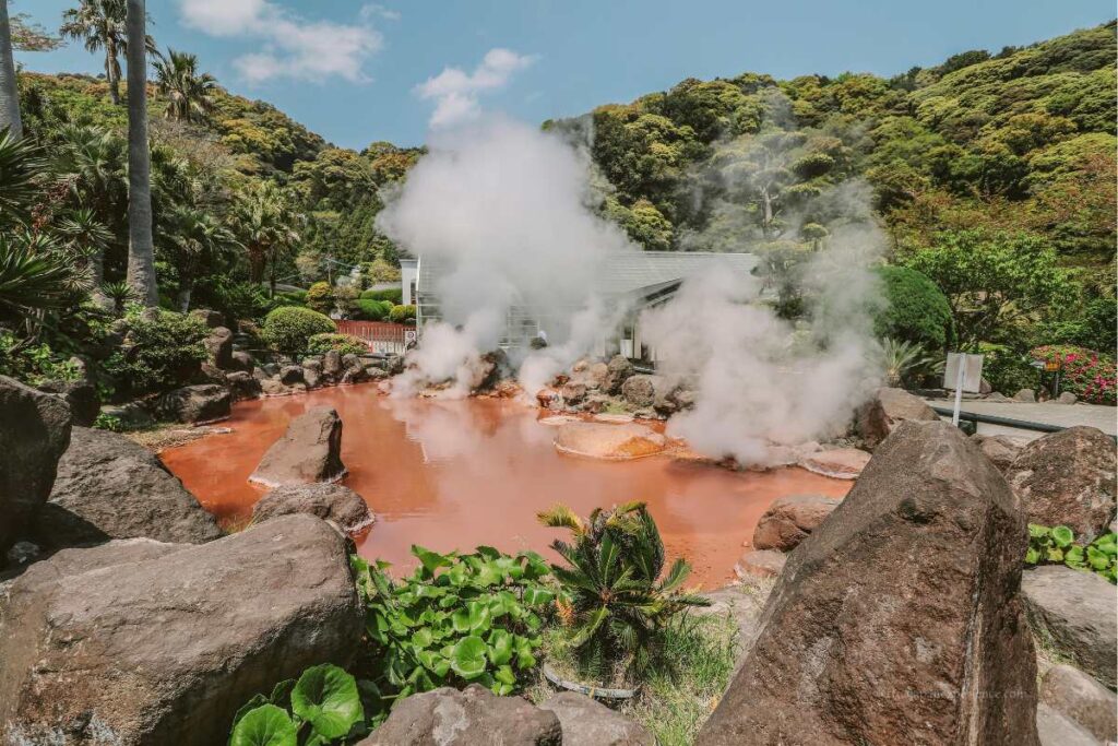 A reddish hot spring pond in Beppu, Japan, surrounded by rocks and tropical greenery, with steam rising from the surface.