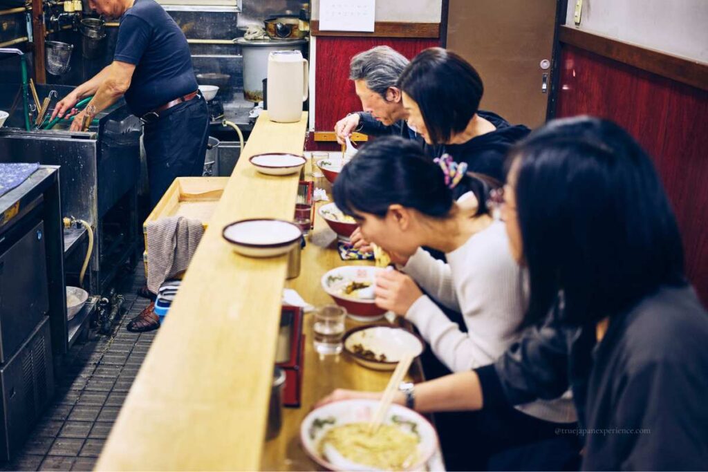 Customers eating ramen at a small Japanese ramen shop, seated at a narrow counter while the chef prepares noodles in the open kitchen.