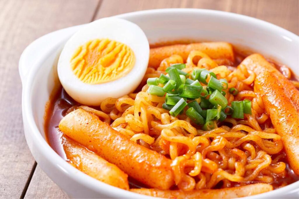 A bowl of spicy Korean ramen with tteokbokki rice cakes, topped with chopped green onions and half a boiled egg.