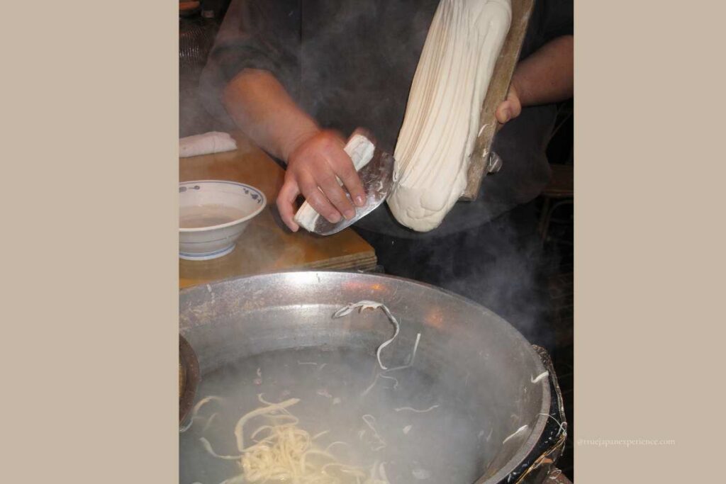 A chef slicing handmade knife-cut noodles into a boiling pot of water, surrounded by steam in a traditional kitchen setting.