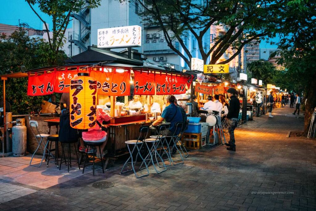 A lively row of Japanese yatai (food stalls) serving ramen and other street food at night, with people sitting and enjoying their meals under red lanterns.