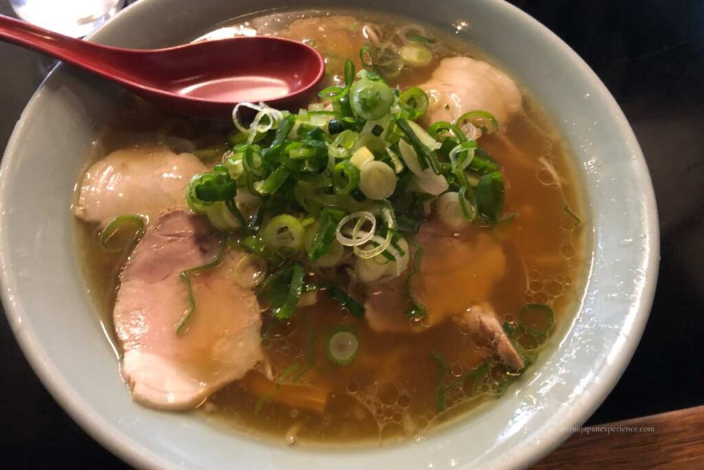 A bowl of soy sauce ramen topped with sliced pork and a generous amount of chopped green onions, served with a red soup spoon.