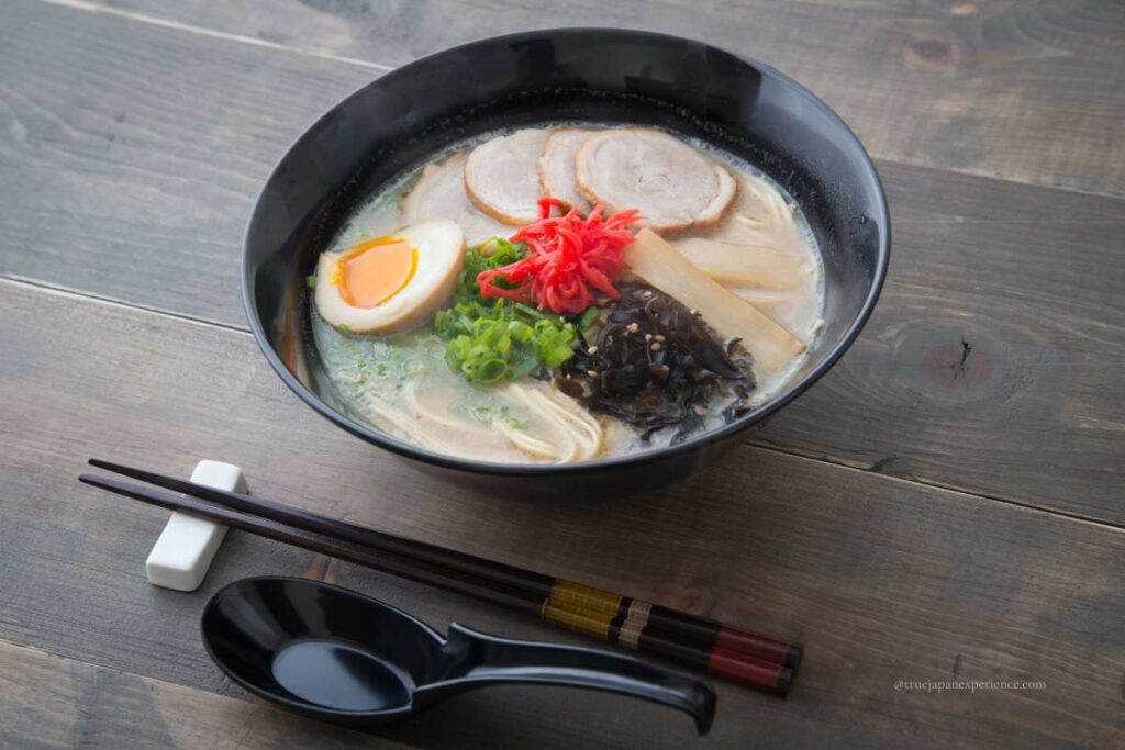 A bowl of tonkotsu ramen served with soft-boiled egg, sliced pork, green onions, pickled ginger, bamboo shoots, and black fungus in a black bowl on a wooden table.
