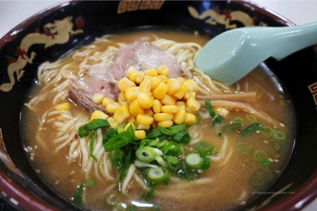 A bowl of miso ramen topped with sweet corn, sliced pork, green onions, and bamboo shoots, served in a black bowl with a dragon design.