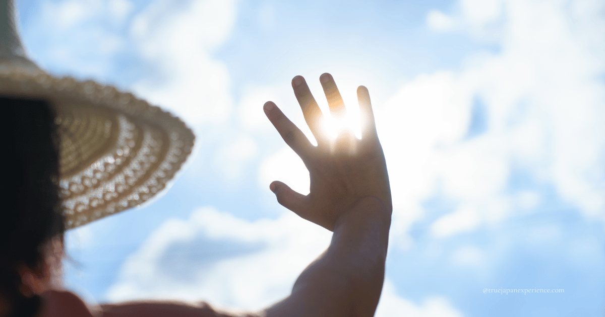 A person in a straw hat shielding their eyes from the strong summer sun with their hand against a bright blue sky — symbolizing the intense heat of Japanese summer.