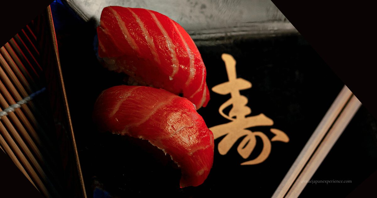 Two pieces of tuna nigiri sushi placed on a black plate with golden kanji for '寿' (kotobuki, meaning longevity or congratulations), accompanied by wooden chopsticks.