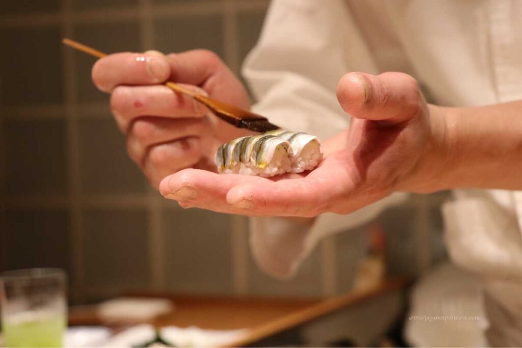 A sushi chef carefully brushing sauce onto a piece of kohada (gizzard shad) nigiri in their hand at a traditional sushi counter.