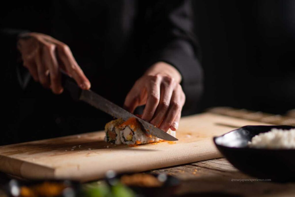 A sushi chef slicing a sushi roll with a sharp knife on a wooden cutting board, with a bowl of rice and ingredients in the background.
