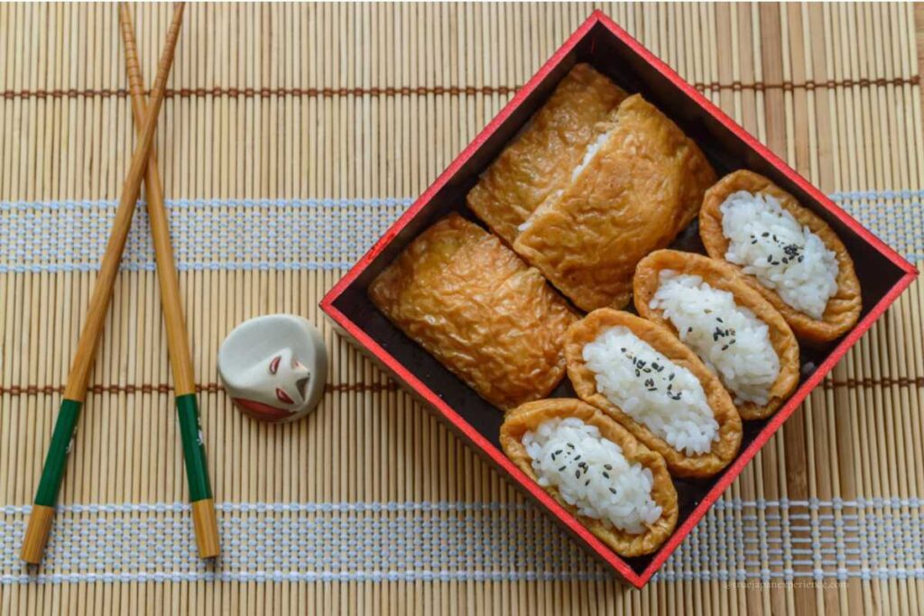 Inari sushi neatly arranged in a red lacquer box, placed on a bamboo mat with chopsticks and a fox-shaped chopstick rest beside it.