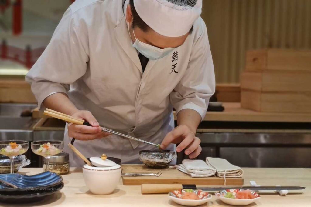 A sushi chef wearing a mask and traditional uniform carefully preparing a dish at the counter, using chopsticks to arrange ingredients with precision.