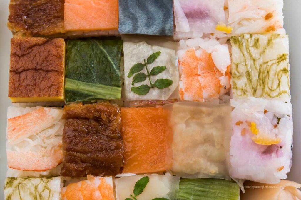 Assorted colorful oshizushi (pressed sushi) arranged neatly in a grid, made with ingredients like shrimp, salmon, egg, vegetables, and seaweed.