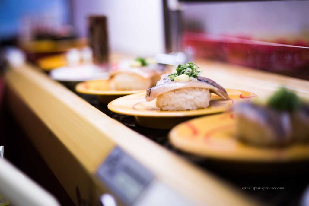 Sushi plates moving along a conveyor belt at a kaitenzushi (conveyor belt sushi) restaurant, with mackerel nigiri topped with green onions and ginger in focus.