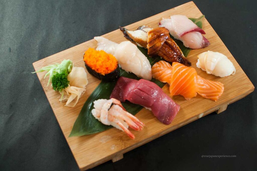 An assorted sushi platter on a wooden board, featuring nigiri with salmon, tuna, shrimp, eel, scallop, and fish roe, served with wasabi and pickled ginger.