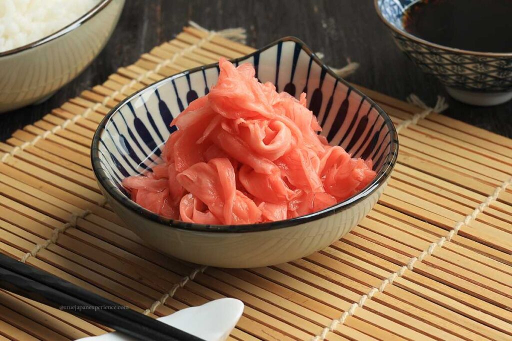 A small bowl of pickled ginger (gari), served on a bamboo mat beside soy sauce and rice, typically enjoyed with sushi.