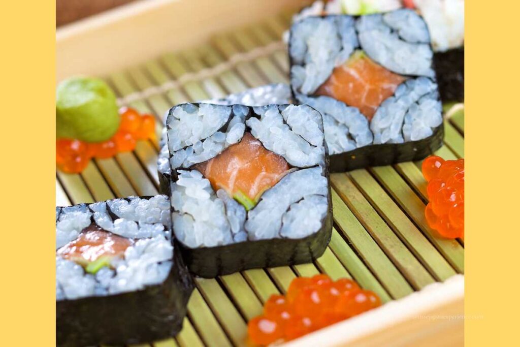 Decorative square makizushi rolls filled with salmon and vegetables, arranged on a bamboo tray with salmon roe and wasabi on the side.