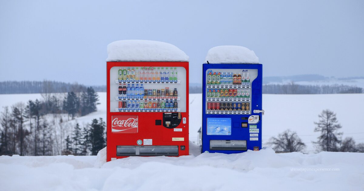 Two colorful Japanese vending machines, one red and one blue, stand side by side in a snowy rural landscape. Snow is piled thickly on top of both machines, with trees and snow-covered fields stretching into the background.