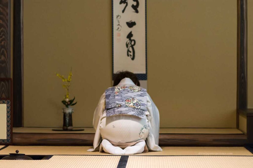A woman in a kimono bowing respectfully in a traditional tea room in Kyoto, with a hanging scroll and seasonal flower arrangement in the background.