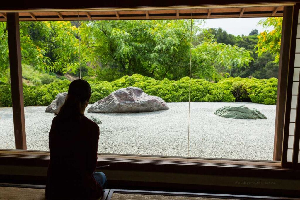 A visitor gazing out at a Zen rock garden from inside a traditional wooden room in Kyoto, framed by lush green trees and carefully raked gravel.