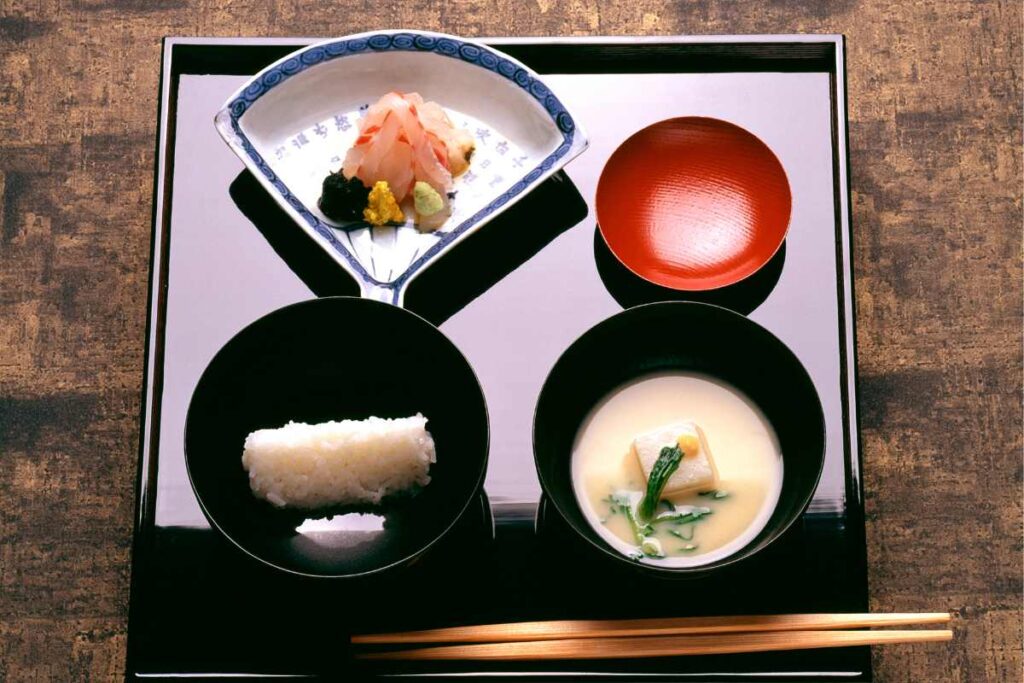 Elegant tray of Kyoto-style Kaiseki cuisine featuring rice, clear soup, tofu in creamy broth, and seasonal sashimi, arranged in lacquerware bowls.