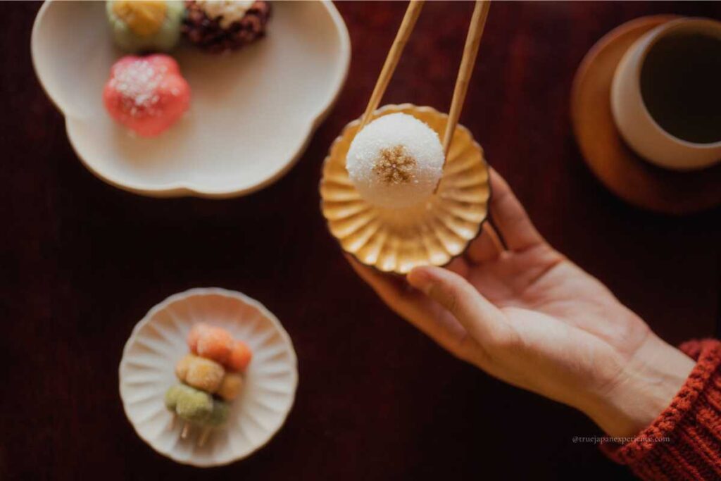 A hand holding a delicate wagashi (Japanese sweet) with chopsticks, surrounded by other colorful sweets and a cup of green tea on a wooden table.
