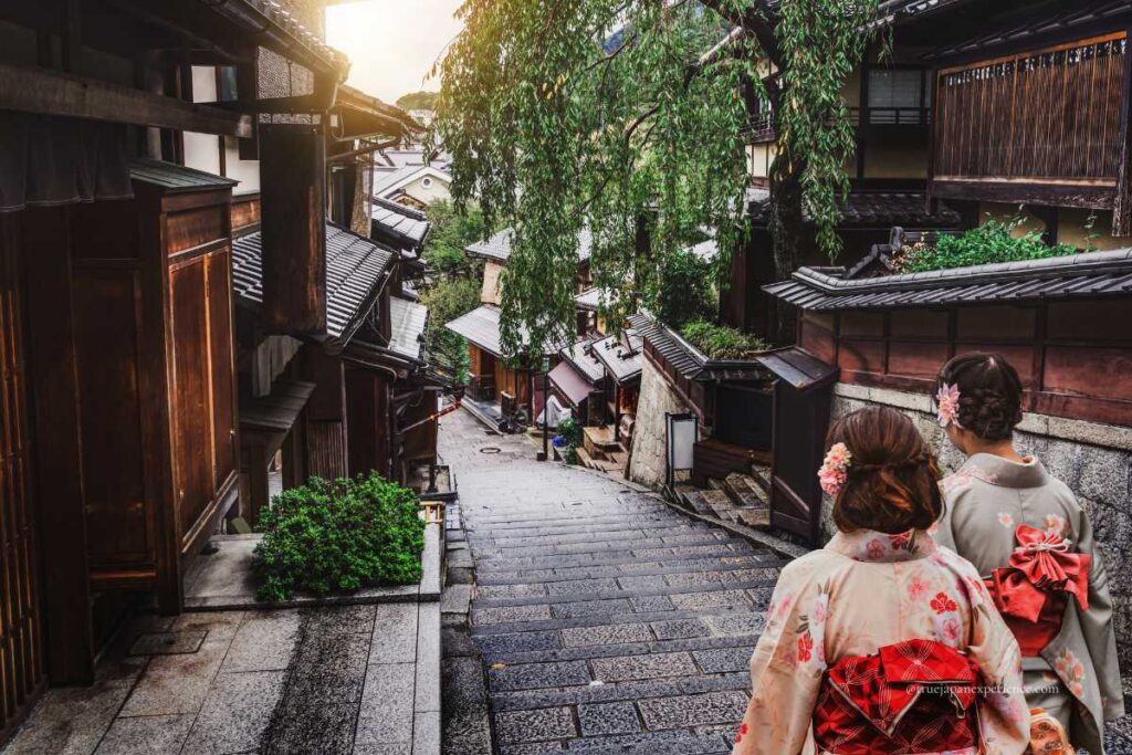 Two women in kimono walking down a stone-paved slope in Kyoto’s historic Higashiyama district, surrounded by traditional wooden buildings and lush greenery.