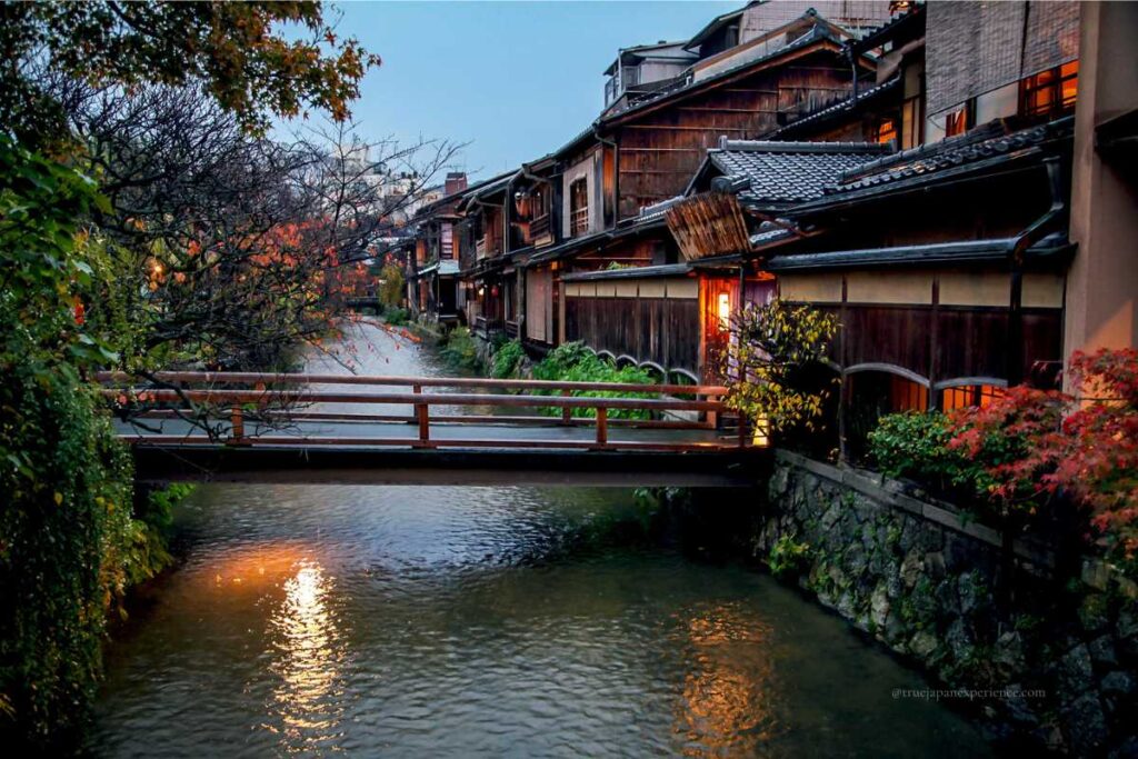 A peaceful twilight view of Shirakawa Canal in Kyoto’s Gion district, with traditional wooden houses, autumn leaves, and a small wooden bridge reflecting in the water.