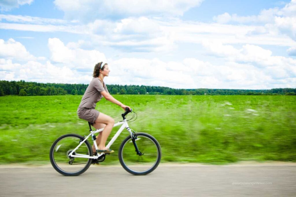 A woman rides a white bicycle along a rural road, passing through green fields under a bright, partly cloudy sky.