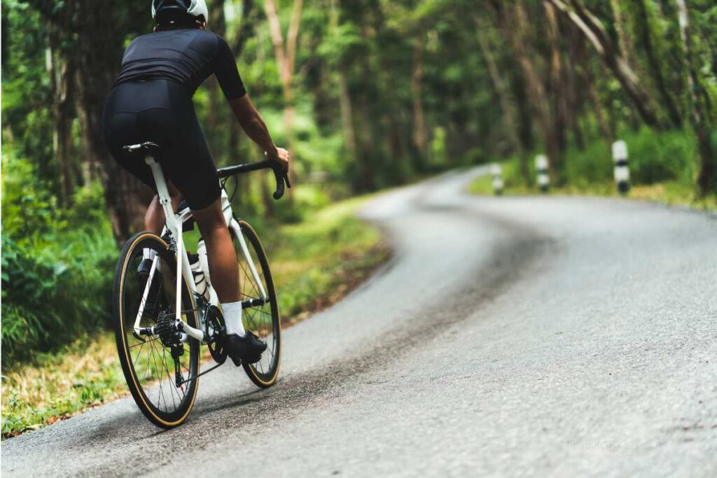 A cyclist in black riding gear climbs a winding forest road on a road bike, surrounded by lush greenery.