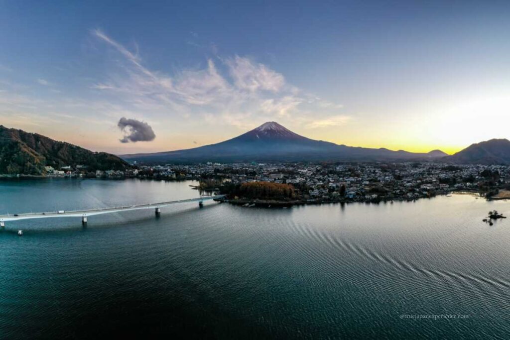 Aerial view of Lake Kawaguchiko and the town below, with Mt. Fuji rising in the distance under a glowing evening sky.