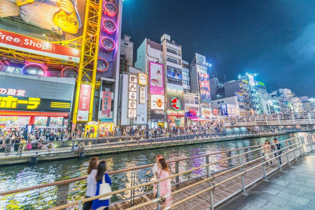 Night view of Dotonbori Canal in Osaka, with colorful neon signs, shops, and people walking along the riverside.
